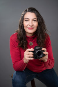 Zuzana Gedeon Headshot in red turtleneck and necklace, holding a camera